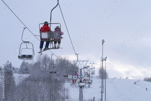 Canvas Print A chairlift with a child and a ski instructor in the chairs at a height in the foreground against the backdrop of the picturesque snow-covered landscape of the ski resort