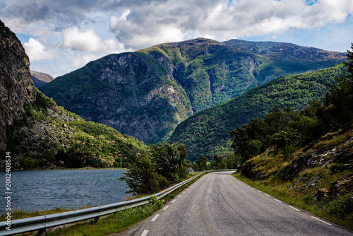 Winding Mountain Road Beside a Serene Lake and Towering Green Peaks Under a Dramatic Sky