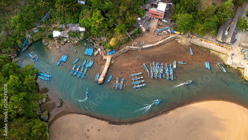 Aerial view of Baron Beach featuring traditional fishing boats moored in the calm bay, surrounded by lush cliffs and sandy shores in Tanjungsari, Special Region of Yogyakarta, Indonesia.