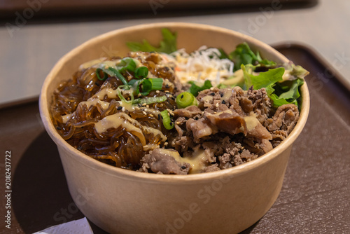 A kraft paper takeout bowl holds Korean-style bulgogi beef and glossy glass noodles topped with green onion, sesame seeds, and a creamy goma sauce, served with fresh salad greens.