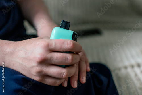 Tablou pe pânză In his hands is a compact gadget - an electronic cigarette or vape in turquoise (mint) color with a black mouthpiece or top panel