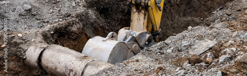 Excavator digging a trench narrow ditch in the dirt to work on underground utilities, municipal construction project, old concrete pipe exposed
