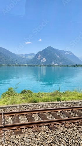 Fotografie A vertical view of a beautiful turquoise alpine lake surrounded by majestic mountains under a clear blue sky, with train tracks running along the shore in the foreground