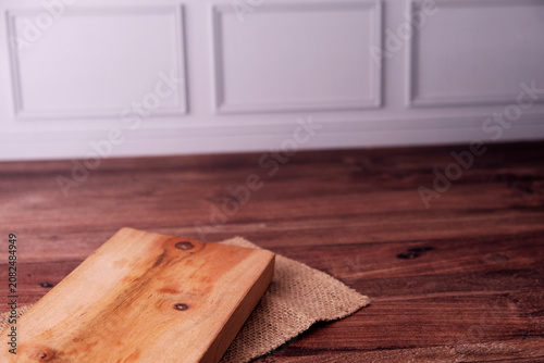 Warm wooden plank rests on burlap atop rich hardwood floor. Soft lighting highlights natural grain and subtle knots. Minimalist setup suggests calm, rustic kitchen or dining space