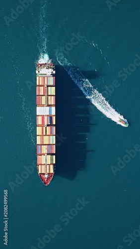 Valokuva Container ship loaded with colorful stacked containers viewed from directly above on open sea as pilot boat departs starboard side