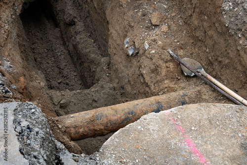 Deep narrow dirt trench with shovel and tamper bar, old iron pipe exposed in underground utilities construction project

