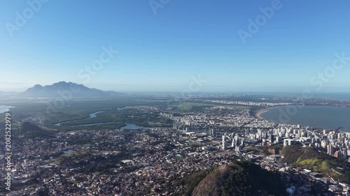 Aerial panoramic view of Vitória, Espírito Santo, Brazil, featuring Vitória Bay, islands, mountains, urban skyline, coastline, and clear blue sky captured by drone.