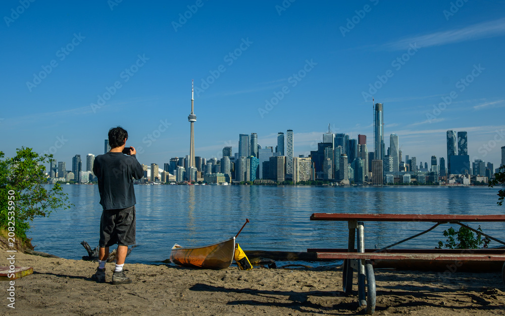 Obraz premium Person photographing the Toronto skyline from a sandy island shore with a canoe on the beach.