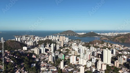 Aerial panoramic view of Vitória, Espírito Santo, Brazil, featuring Vitória Bay, islands, mountains, urban skyline, coastline, and clear blue sky captured by drone.