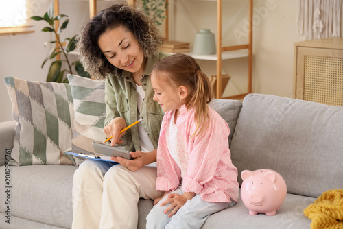 Little girl with her mother, calculator and piggy bank sitting on sofa at home