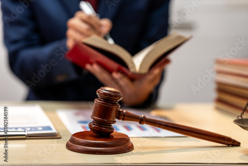 Wooden gavel on a desk with a person writing in a book in the background, showing a legal office scene with law and justice concept.