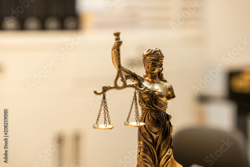 Close view of a bronze lady justice statue with balanced scales in a softly blurred office or courtroom setting, symbolizing law, fairness, order, and legal services.