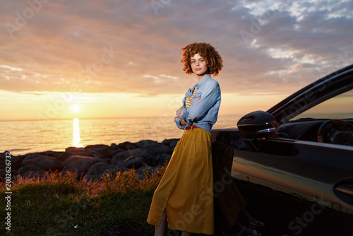 Foto Curly haired woman posing beside a car at sunset, wearing a denim jacket and mustard skirt, overlooking a rocky coastline
