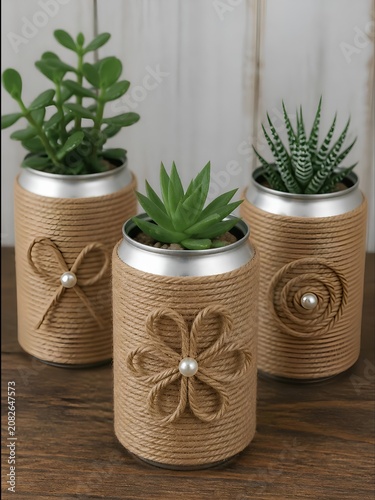 Three miniature plants, including succulents and a cactus, are creatively potted in repurposed aluminum cans. The cans are wrapped in natural jute twine, adorned with twine designs resembling flowers.