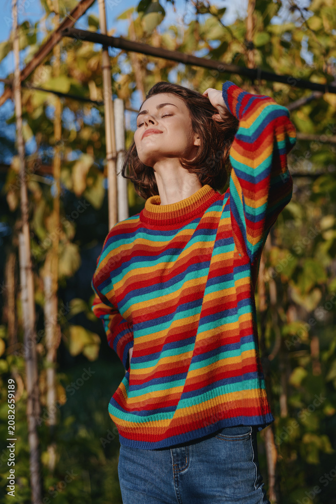 Fototapeta premium Woman in a colorful striped sweater outdoors among vines, relaxed and smiling with short hair, casual outfit, hand behind head, warm golden hour lifestyle portrait.