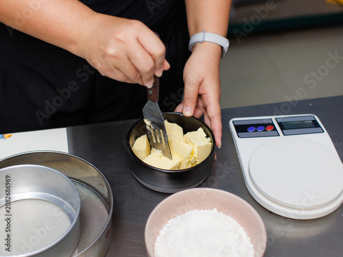 Asian woman preparing ingredients scooping butter into bowl weighing it kitchen digital scale mixing flour cocoa powder sugar make delicious homemade brownies dessert cake recipe baking process bakery