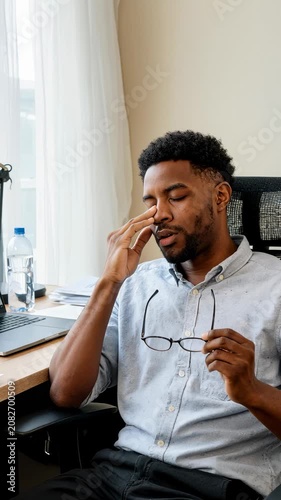 Man sits at desk, rubbing tired eyes. He holds glasses, looks exhausted. Office setting, natural light from window. Suggests burnout, stress, or work fatigue