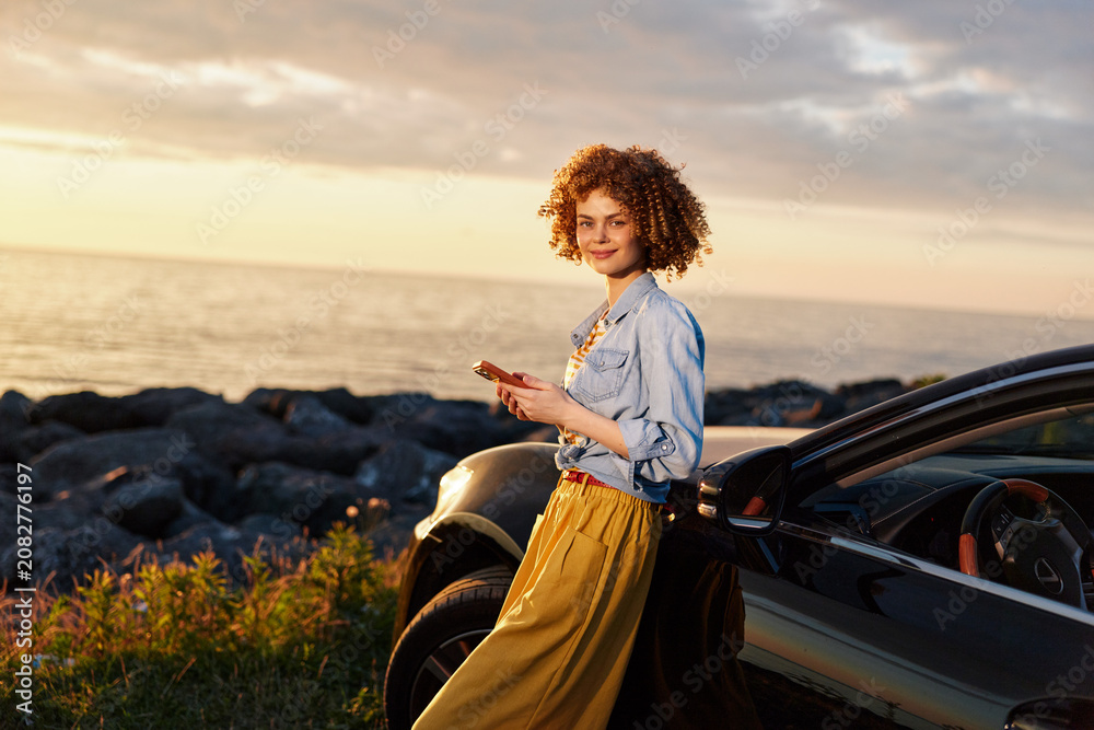 Fototapeta premium Cheerful woman outdoors by a car at sunset, wearing a denim shirt and mustard skirt, enjoying a relaxed lifestyle with coastal breeze and warm tones and golden light on her smile