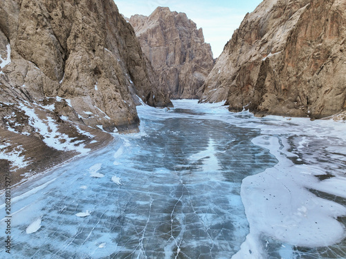 Wide aerial view looking down a narrow mountain gorge, filled with expansive, fractured blue ice between towering, textured rock walls with snow patches