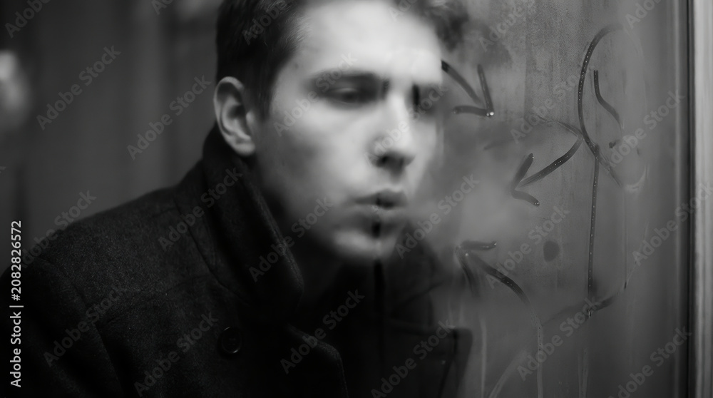 Fototapeta premium Black and white cinematic portrait of a man exhaling smoke near a window, evoking introspection, melancholy, mystery, and vintage noir atmosphere.
