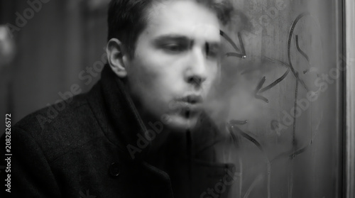 Black and white cinematic portrait of a man exhaling smoke near a window, evoking introspection, melancholy, mystery, and vintage noir atmosphere.