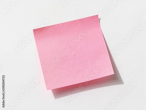 A single square pink sticky note placed on a smooth white background with gentle directional lighting. The paper shows subtle texture and a slightly lifted