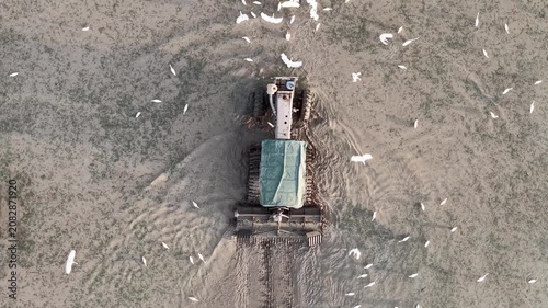 Aerial view of tractor tilling soil on farm field surrounded by birds flying