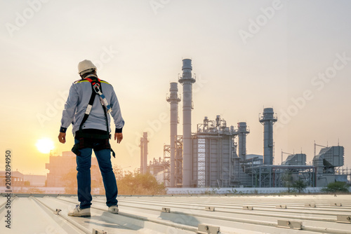 Foto Industrial Sunrise: A lone worker, outfitted with safety gear, stands on a rooftop, surveying a vast industrial complex as the sun rises, casting a warm golden hue over the scene