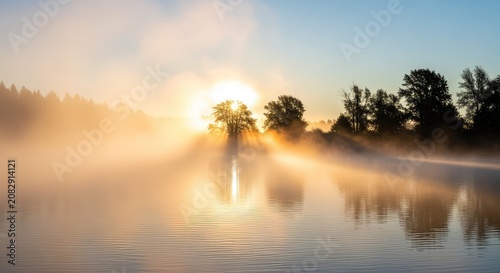 Dense white fog rolls over a calm river at dawn as the sun rises, casting soft light through the trees and reflecting on the still water, scenic, dreamy, soft