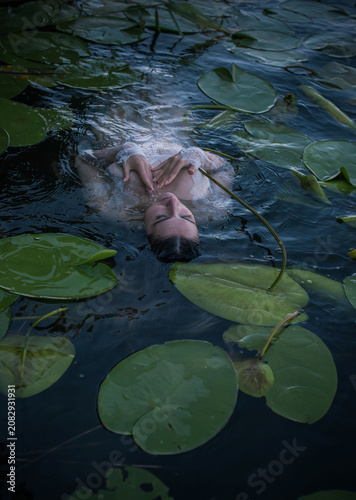 Mystical Slavic mermaid floating in deep lake water among green water lilies, pagan nymph undine in white lace dress, dark fairytale and mythology concept