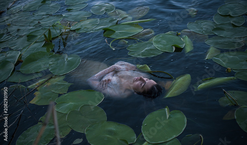Fotografie Mystical Slavic mermaid floating in deep lake water among green water lilies, pa