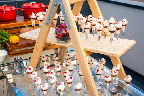 Elegant catering dessert display on a wooden ladder stand, featuring small glass cups with layered trifles topped with cream and edible flowers, perfect for weddings and luxury outdoor events.