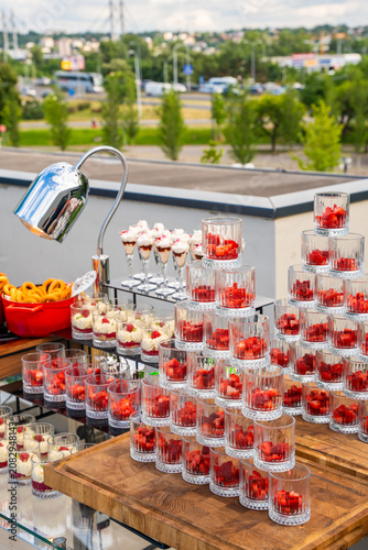 Elegant catering dessert station outdoors with a pyramid of strawberry cups, cream trifles, and snacks on wooden boards, perfect for an upscale event or wedding celebration.