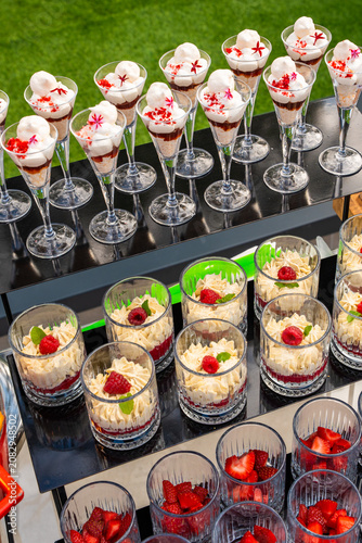 High-angle view of a professional catering dessert display featuring assorted glass cups with creamy trifles, fresh raspberries, meringue toppings, and sliced strawberries for an upscale event.