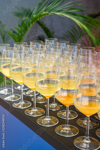 Array of elegant wine glasses filled with refreshing yellow and orange cocktails, arranged on a dark service table at an upscale outdoor event with tropical greenery in the background.