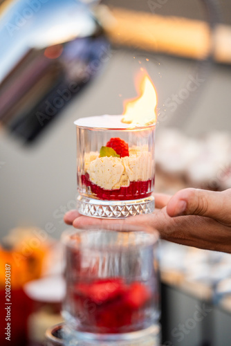 A chef's hand holding a glass cup with a flaming layered dessert, featuring cream, raspberry compote, and fresh fruit at an exclusive outdoor catering event.