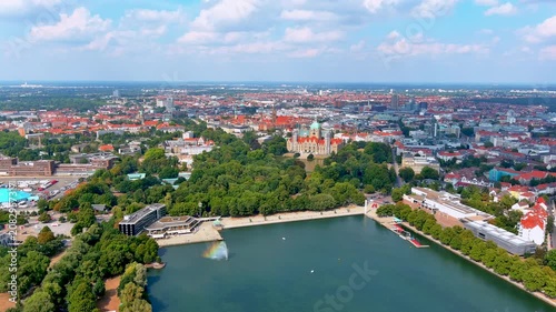 Aerial drone view of Hannover with the New Town Hall, Maschpark, the Maschsee lake fountain, and red rooftops of the city centre, Germany, summer daytime.