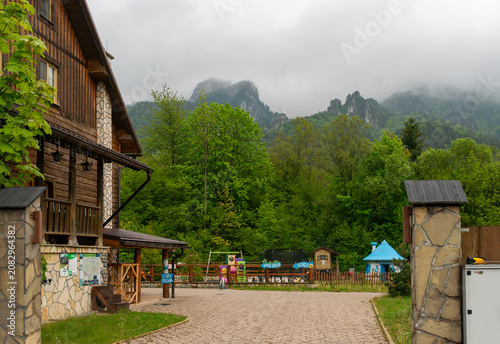 Konstfotografi Wooden guesthouse and stone wall stand below misty limestone cliffs and green forested hills, with playground signs and rural tourism buildings