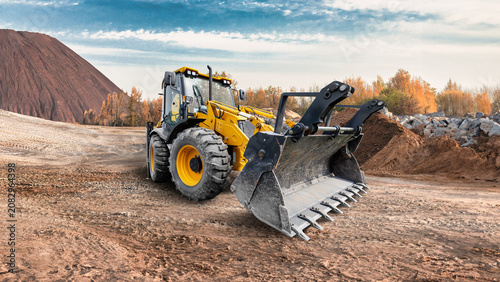 A universal excavator loader operates on a construction site. The machine is moving dirt and rocks in an open area during the day. Trees are visible in the background