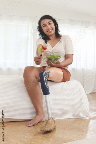 Happy woman with prosthetic leg eating vegetables salad on white bed. Smiling disabled Asian female holds salad bowl, rests in bedroom. Cheerful woman with a disability who uses a prosthetic leg.
