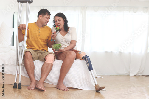 Happy Asian couple spends time together at home, woman with prosthetic leg, man with knee injury eat vegetables salad in bed. Disability lovers hold salad bowl, show love to each other in bedroom.
