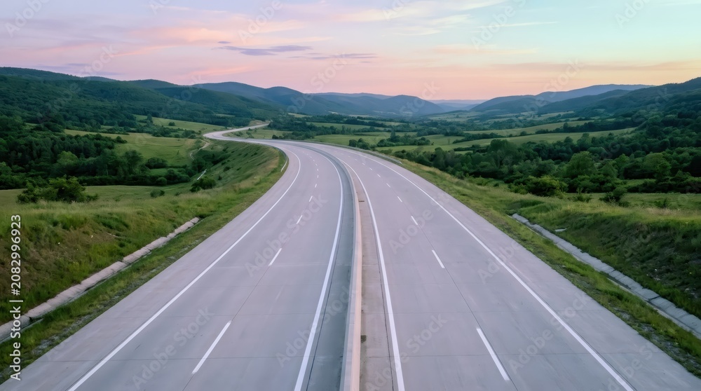 Obraz premium Empty multi-lane highway stretching through summer mountain landscape. Scenic asphalt road winding into distance at sunset. Infrastructure journey concept. Outdoor travel background.