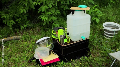 Portable outdoor washing station setup for camping activity, featuring large water container, various cooking utensils, bowls, cups, and soap, surrounded by green grass and foliage.