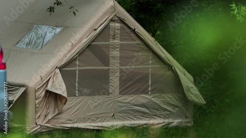 Large beige camping tent with windows and roof vent set up on green grass at edge of forest, suggesting outdoor recreation and temporary shelter in nature