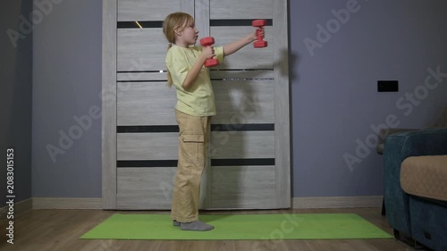 Young caucasian girl performing arm exercises with red dumbbells on green mat indoors. Fitness routine of child for healthy lifestyle at home.