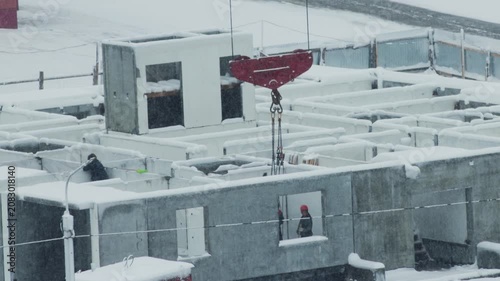 Construction site activity during heavy snowfall, crane moving concrete building section, worker observing progress amid winter conditions
