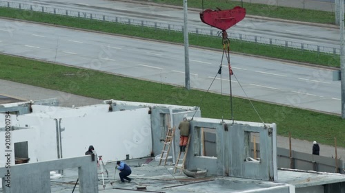 Construction workers assemble pre-fabricated concrete wall panels on building site using large red crane hook overhead by busy road with cars