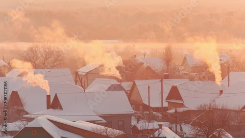 Winter village scene with smoke plumes rising from chimneys, snow-covered rooftops bathed in golden light of frosty morning sunrise.