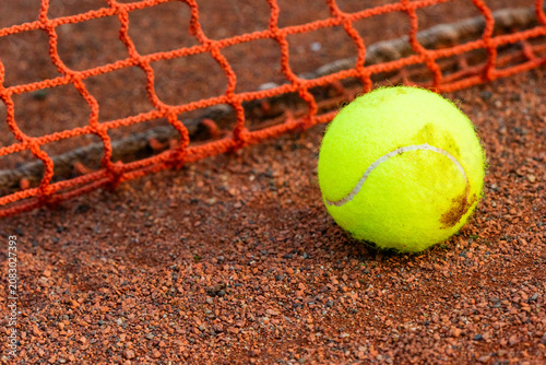 Close-up of yellow tennis ball on clay court with net and blurred background, illustrating tennis season, sport, competition and summer fitness.