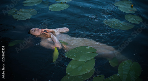 Ethereal woman floating in dark lake water among green lily pads, mystical water nymph or goddess in white shirt, fine art fantasy concept